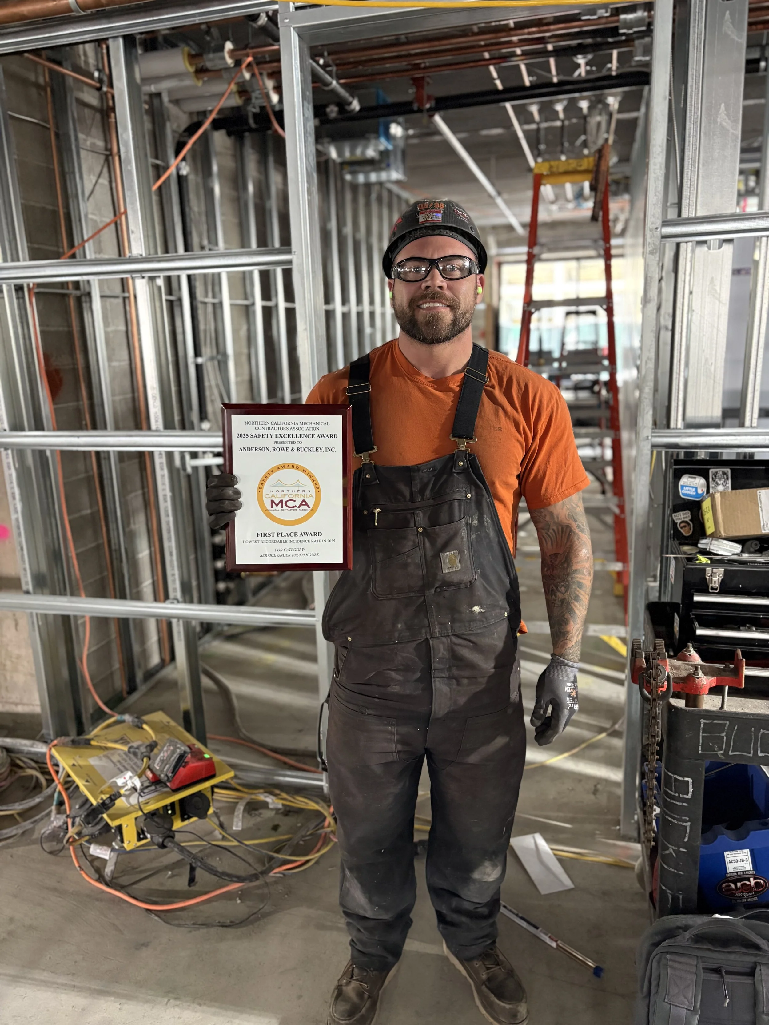 Man in construction attire holding an award plaque at a construction site with metal framing, a red ladder, and construction tools and equipment around him.