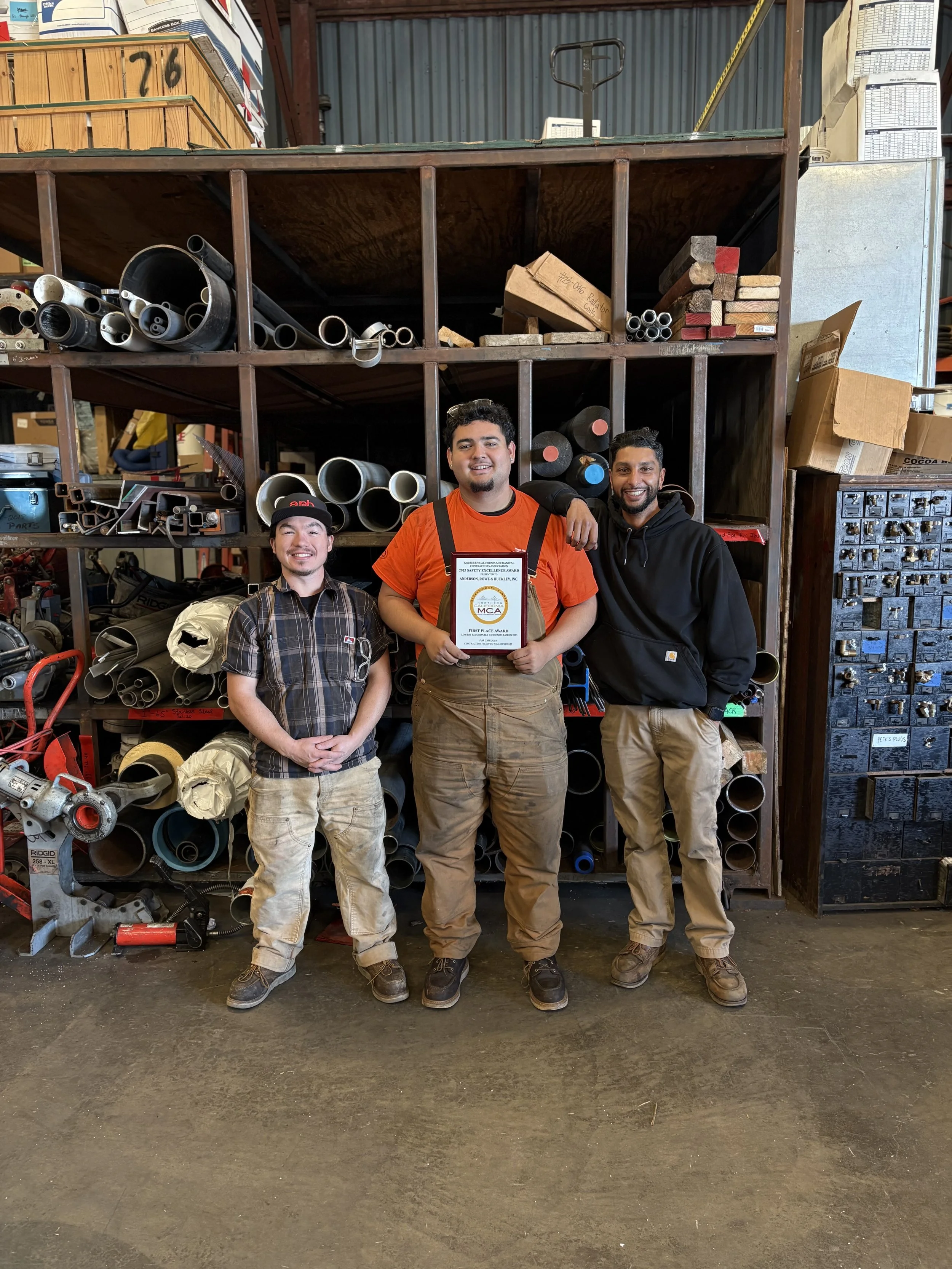 Three young men standing in front of a large shelf filled with metal pipes and equipment in a workshop or warehouse. One man holds a plaque or certificate. They are smiling and dressed in casual work attire.