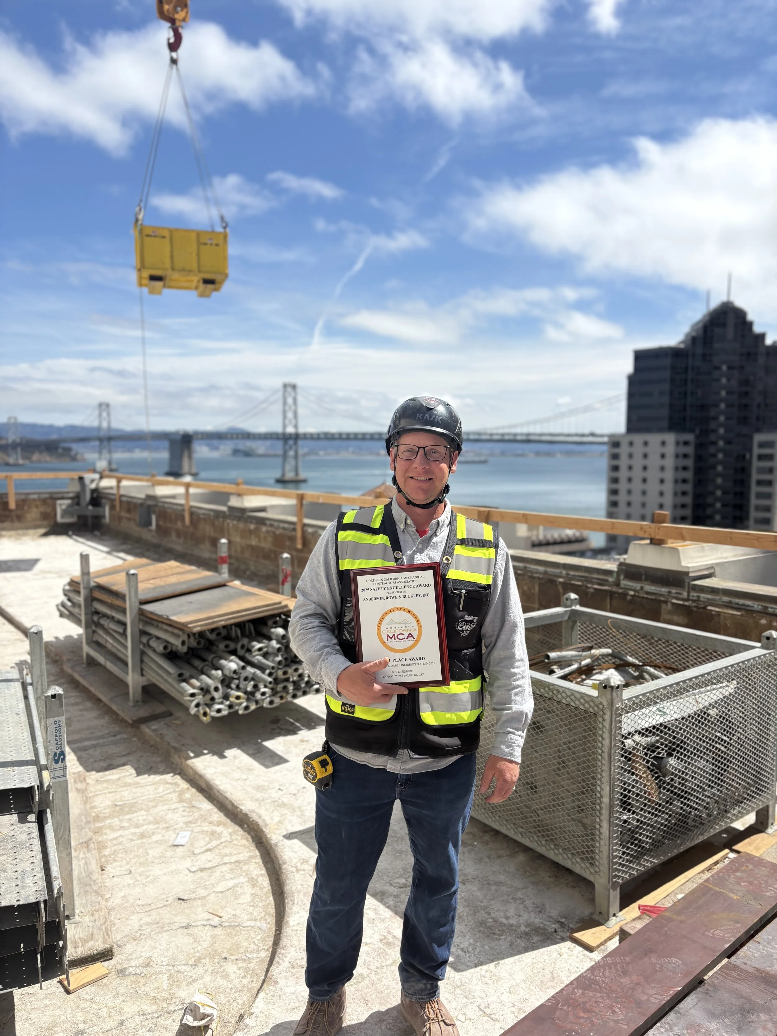 Man in safety vest and helmet holding an award certificate on a construction site with city buildings and bridge in background.