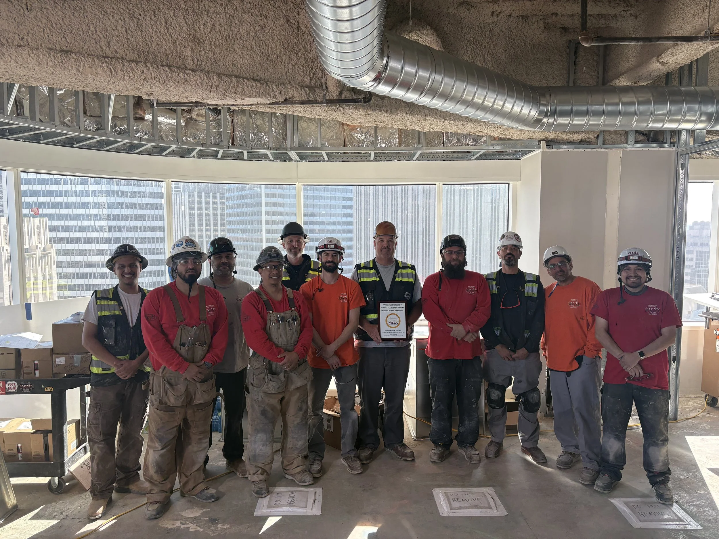 Group of construction workers wearing safety gear and helmets standing inside a building under construction, with large windows and cityscape visible in the background, holding a certificate or award.