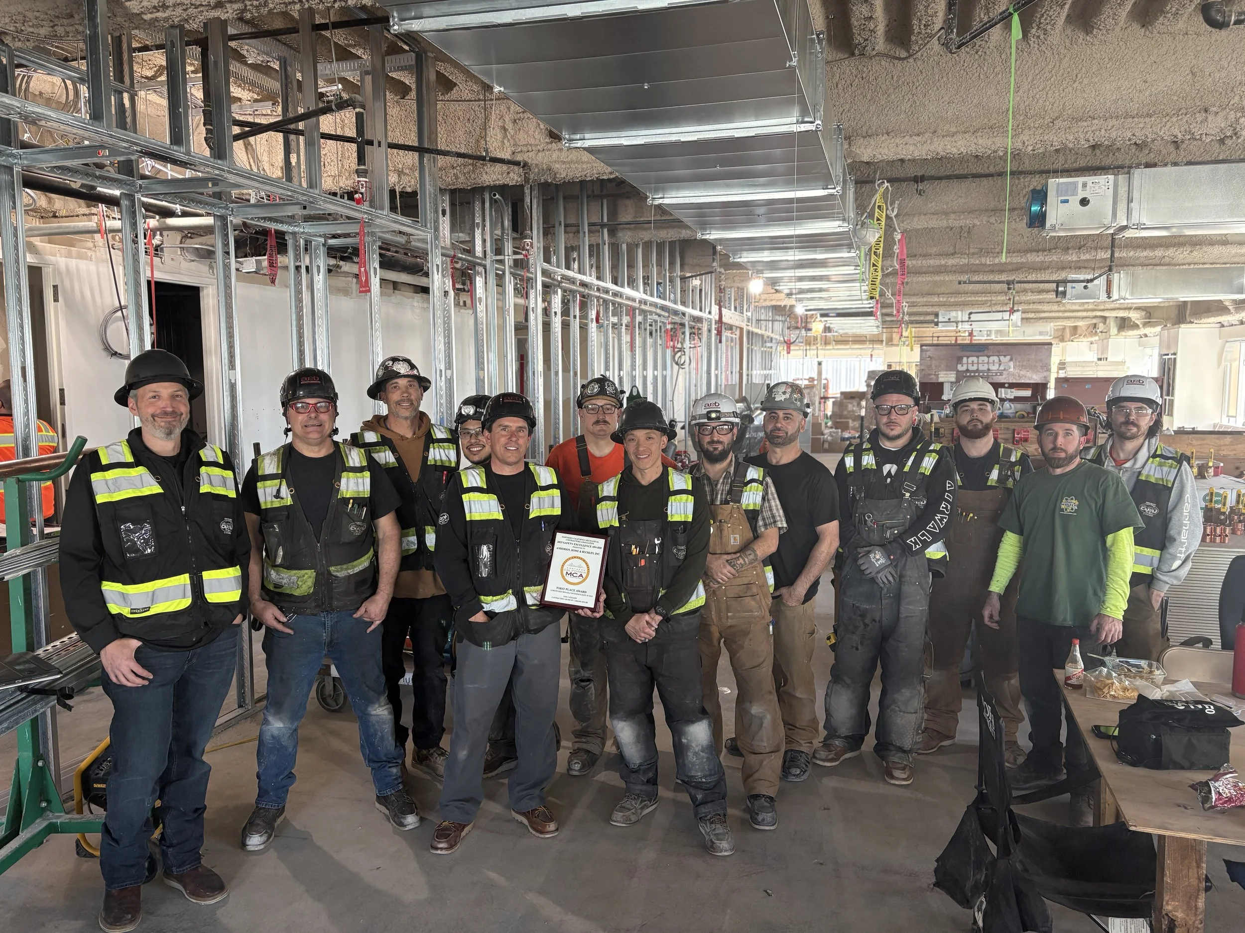 A group of construction workers and managers wearing helmets and safety vests standing inside a building under construction with exposed framework, ductwork, and construction equipment.
