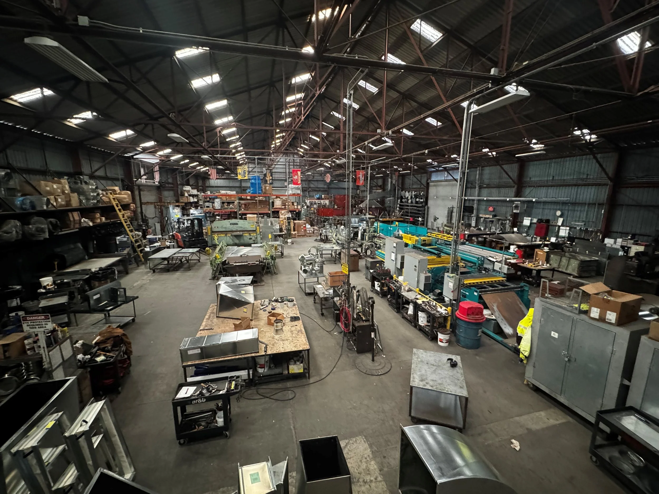 Interior of a large industrial warehouse with high metal roof and various equipment, tools, and machinery scattered throughout the space.