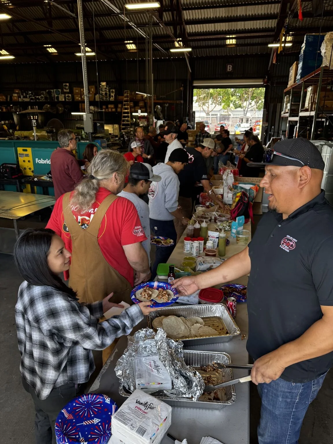 People in a warehouse gathering for a community meal, serving food from trays and bottles with a line of people waiting to receive food.