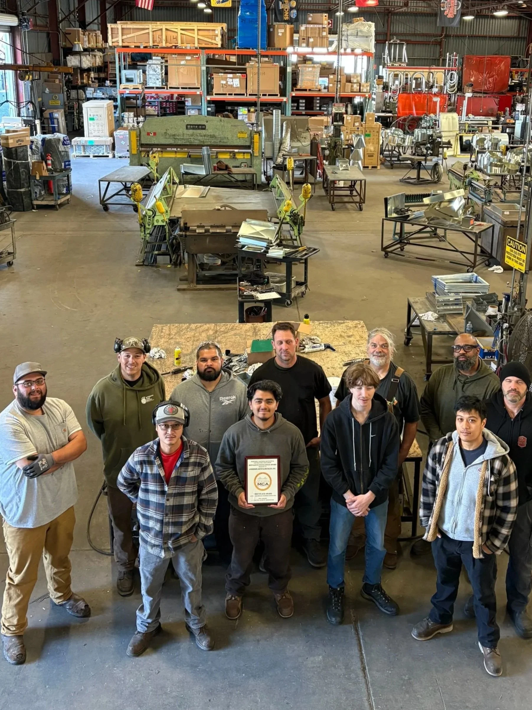 Group of eight people standing in a warehouse, with one person holding a plaque. The warehouse has industrial equipment, shelves with boxes, and work tables in the background.