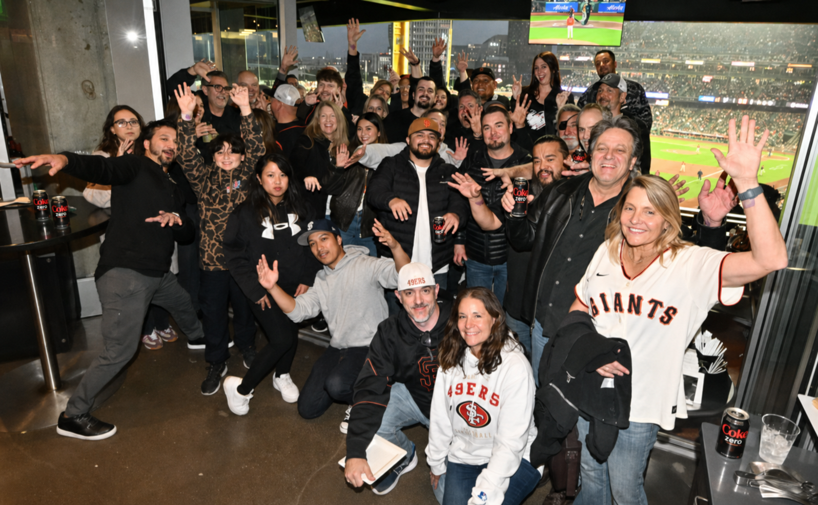 A group of people celebrating and posing for a photo in a sports bar with a view of a baseball game on multiple screens behind them.