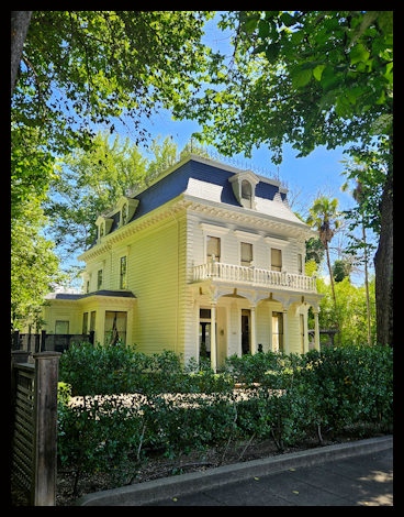 A large, ornate, white Victorian-style house with decorative trim and a steep roof, surrounded by trees and greenery under a clear blue sky.