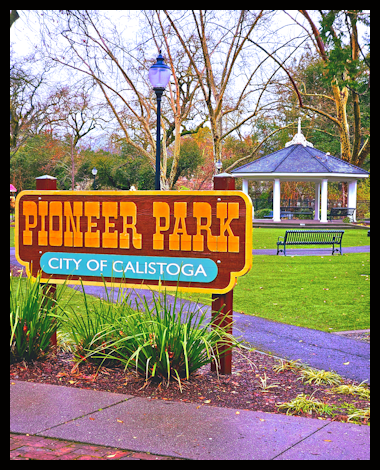 Pioneer Park sign in Calistoga, with a gazebo, benches, lamp posts, trees, and a walking path in the background.