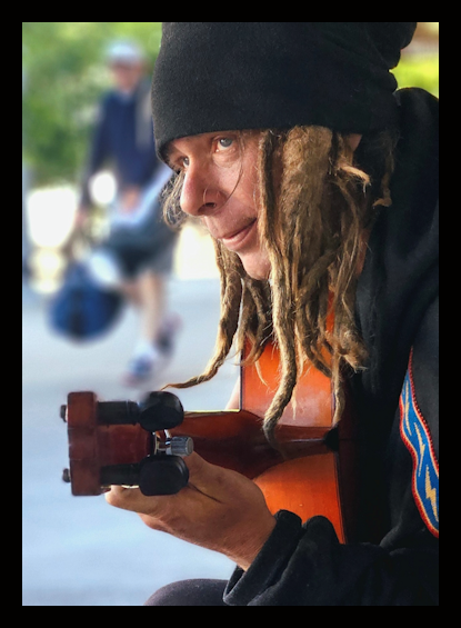 Woman with dreadlocks wearing a black beanie playing guitar outdoors, with blurred figures in background.