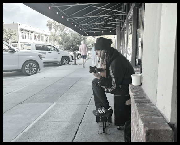 A person with dreadlocks, wearing a black hoodie and beanie, sitting on a small stool outside a storefront, holding a camera, with a cup nearby. Vehicles and pedestrians are visible on the street.