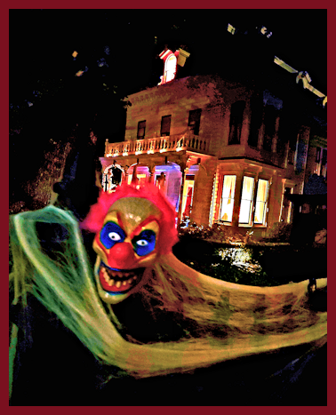 A creepy clown with yellow hair, blue eyes, and a red nose is smiling eerily in front of a lit-up house at night.