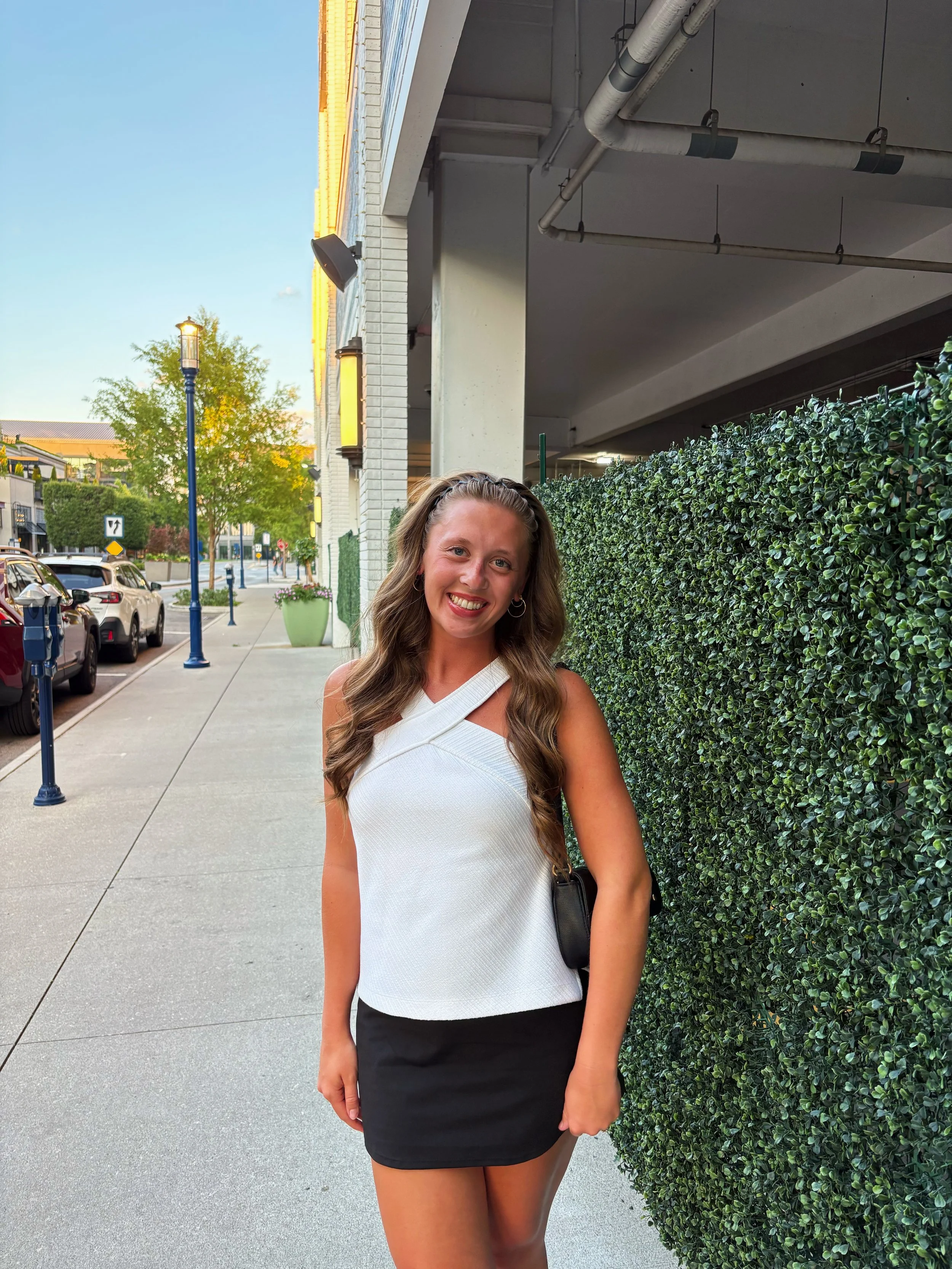 A smiling young woman with long wavy hair in a white sleeveless top and black skirt standing outdoors on a sidewalk next to a green hedge with a city street, cars, trees, and buildings in the background during the daytime.