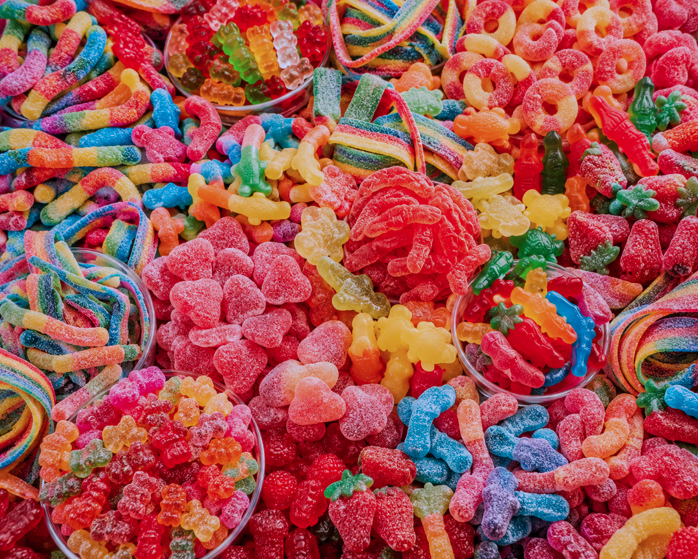 Assorted colorful gummy candies including worms, bears, rings, and sour strips arranged in bowls and scattered on a surface.