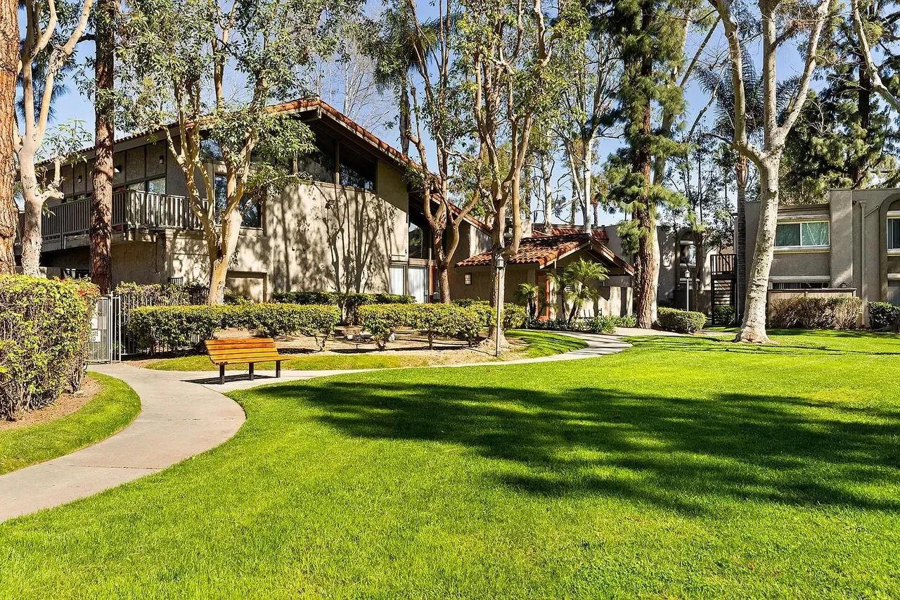 A well-maintained grassy park area with a curved concrete pathway, surrounded by trees, bushes, and residential buildings in the background.