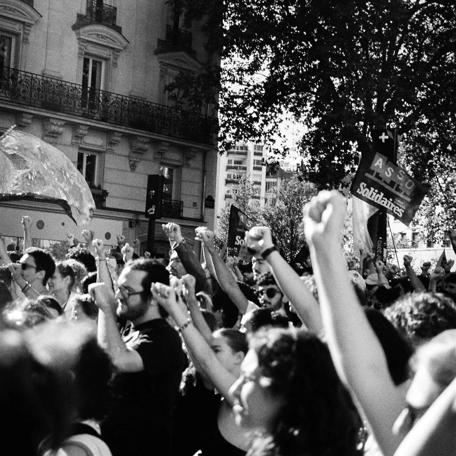 Crowd of people at a demonstration or protest, raising fists and holding flags with the words "ASSO Solidarity" on them, in an urban city street with buildings and trees in the background.