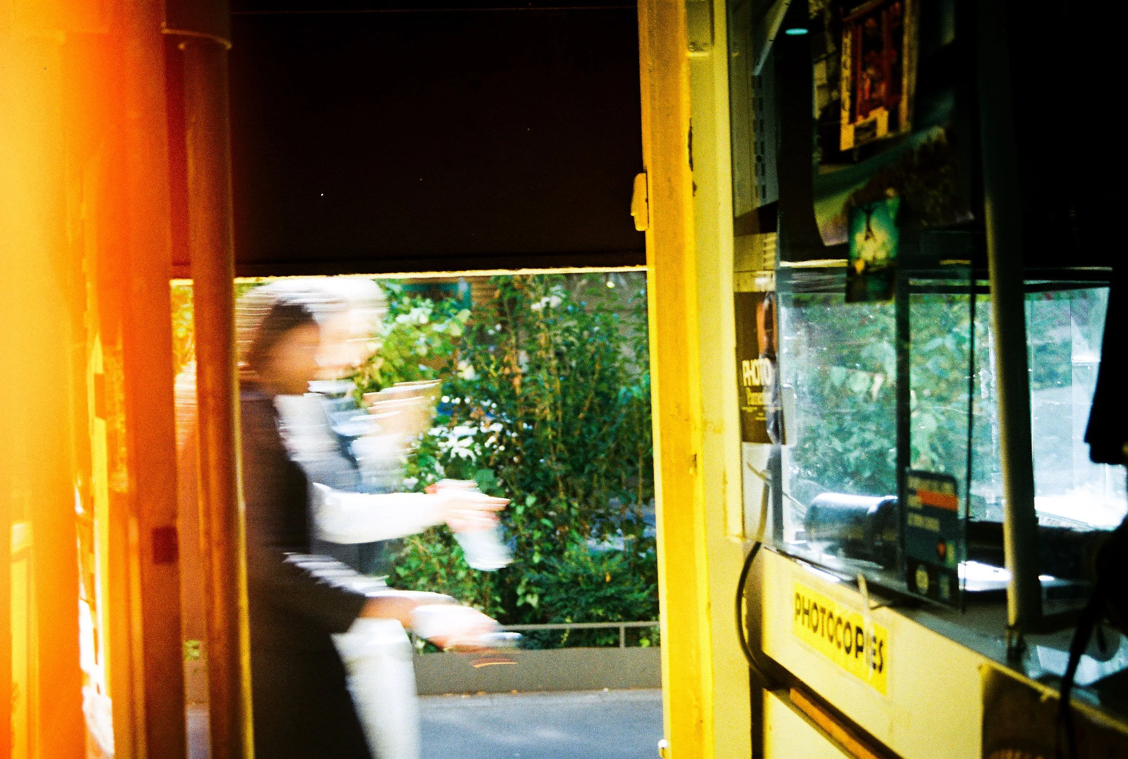 Inside a yellow photo booth with a blurred person holding a white cup, and greenery outside the window.