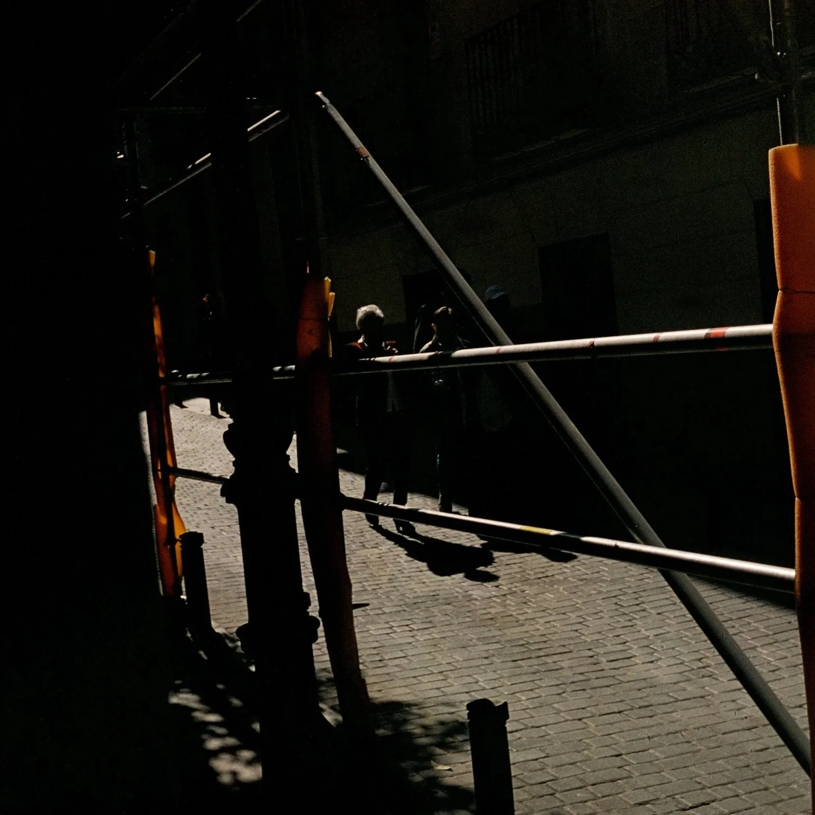 Dimly lit scene of a cobblestone street with scaffolding and construction barriers. Three silhouetted people are standing near the barriers, with shadows cast on the ground. The background is dark, with minimal lighting.