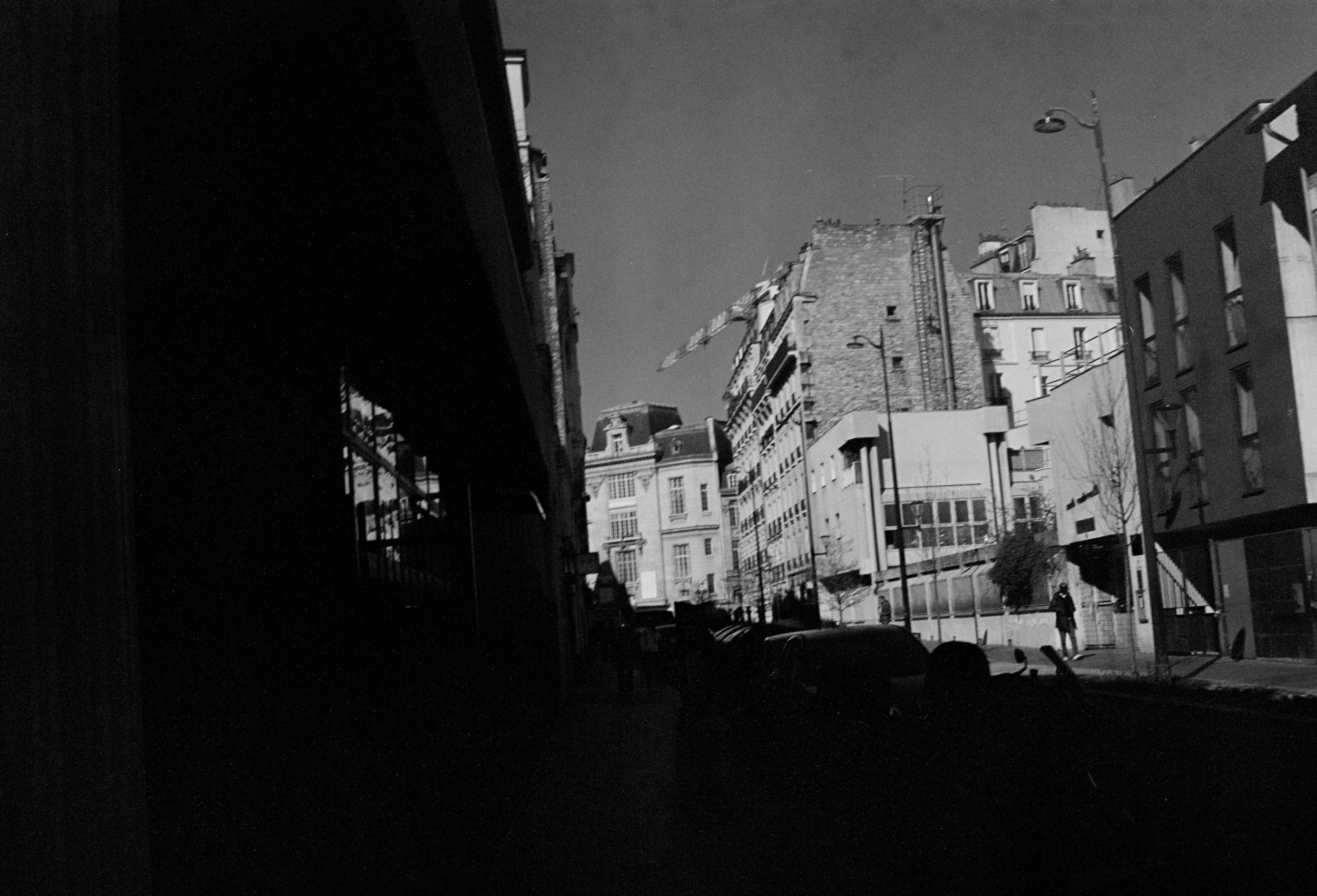A black and white photo of a city street viewed from beneath a bridge or overpass. Buildings with varied architectural styles line the street, and a crane is visible in the background.