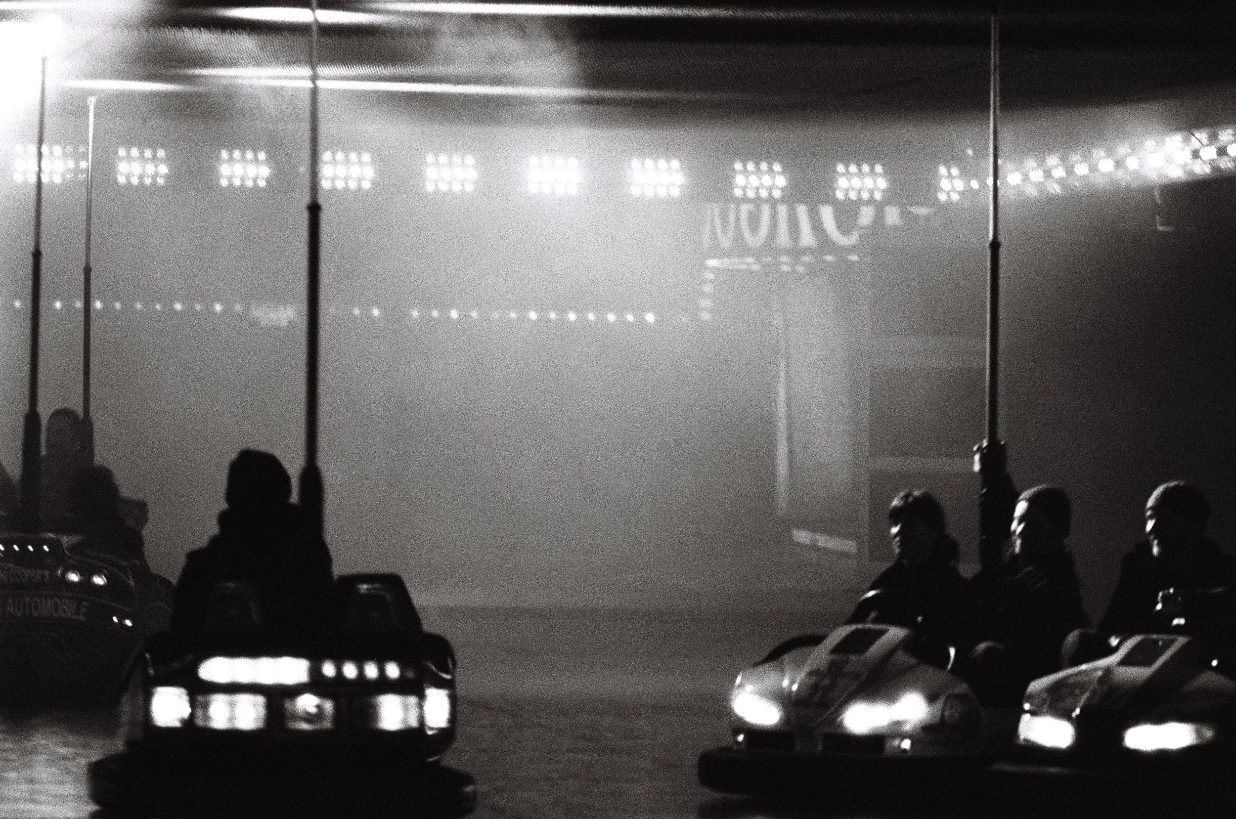 A black and white photo of a parking lot with several cars and people standing near street lamps. Bright lights and reflections are visible in the background.