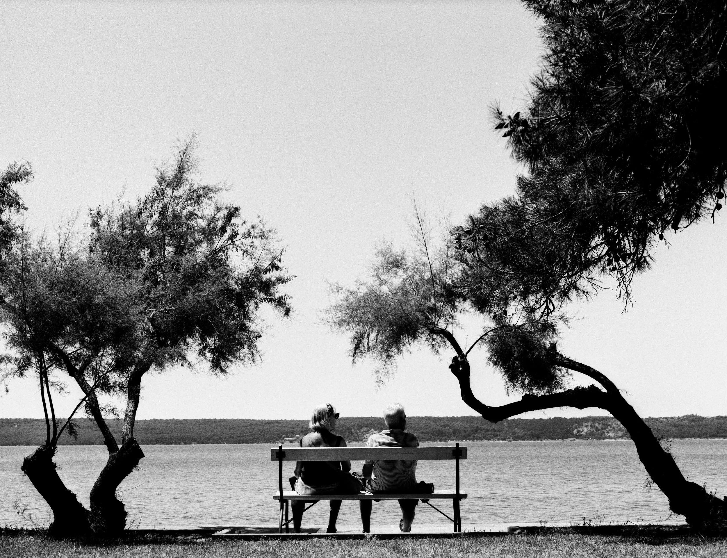 Black and white photo of an elderly couple sitting close together on a bench by the river, framed by trees, with the water and shoreline in the background.