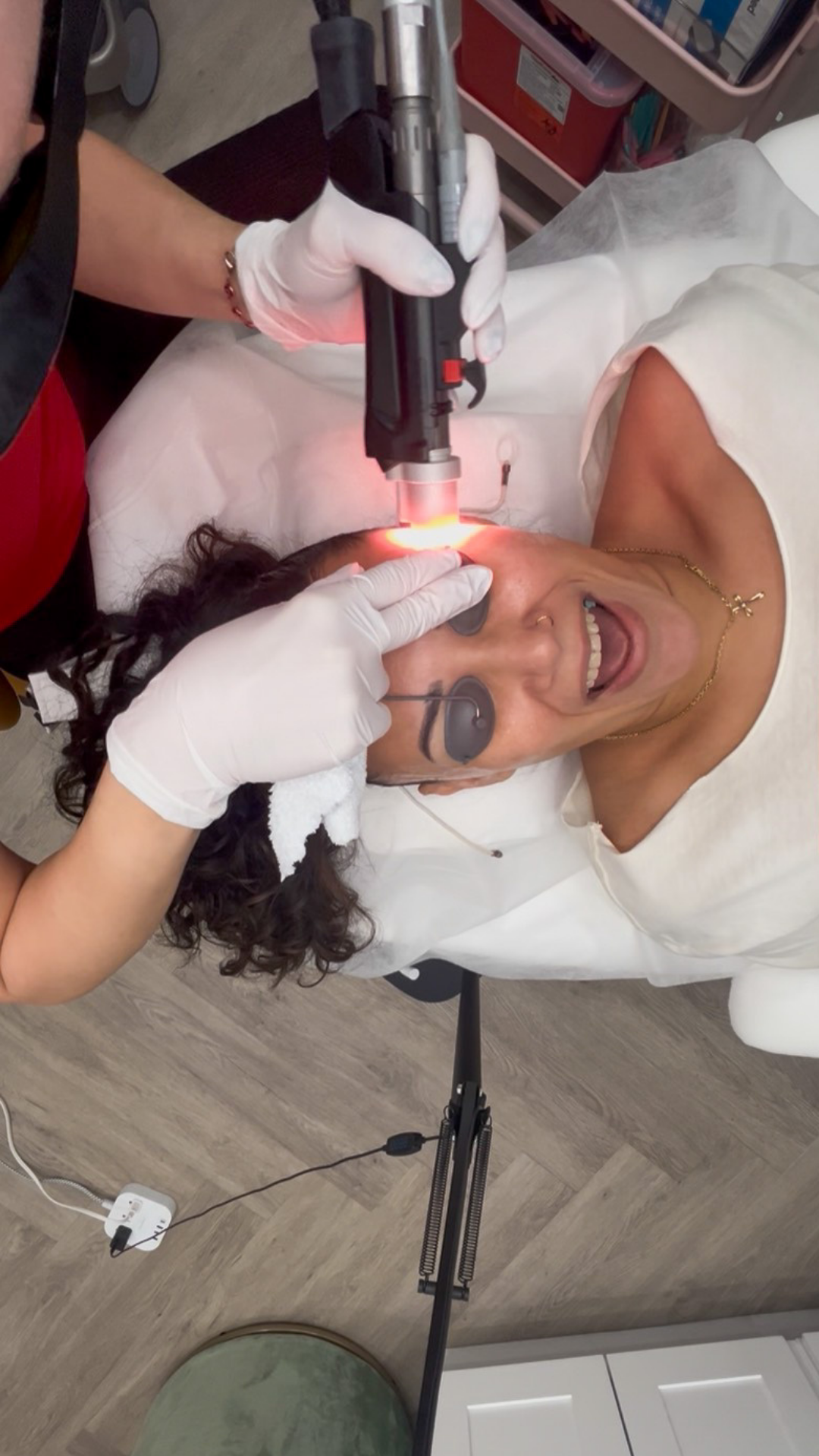 A woman smiling and lying on a treatment chair receiving a Moveo PL with the DEKA Motus AZ+ device on her face from a medical professional wearing gloves.