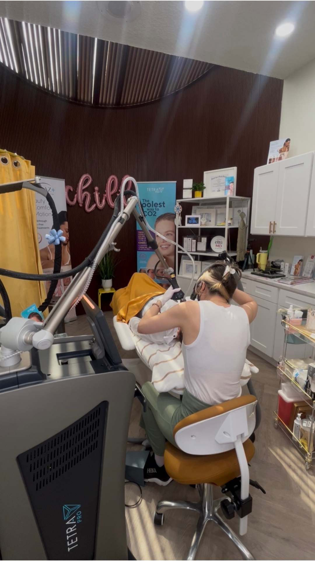 A person lying on a treatment bed receiving DEKA pulse by the same woman with split hair. The clinic has medical laserposters, white cabinets, and medical equipment.