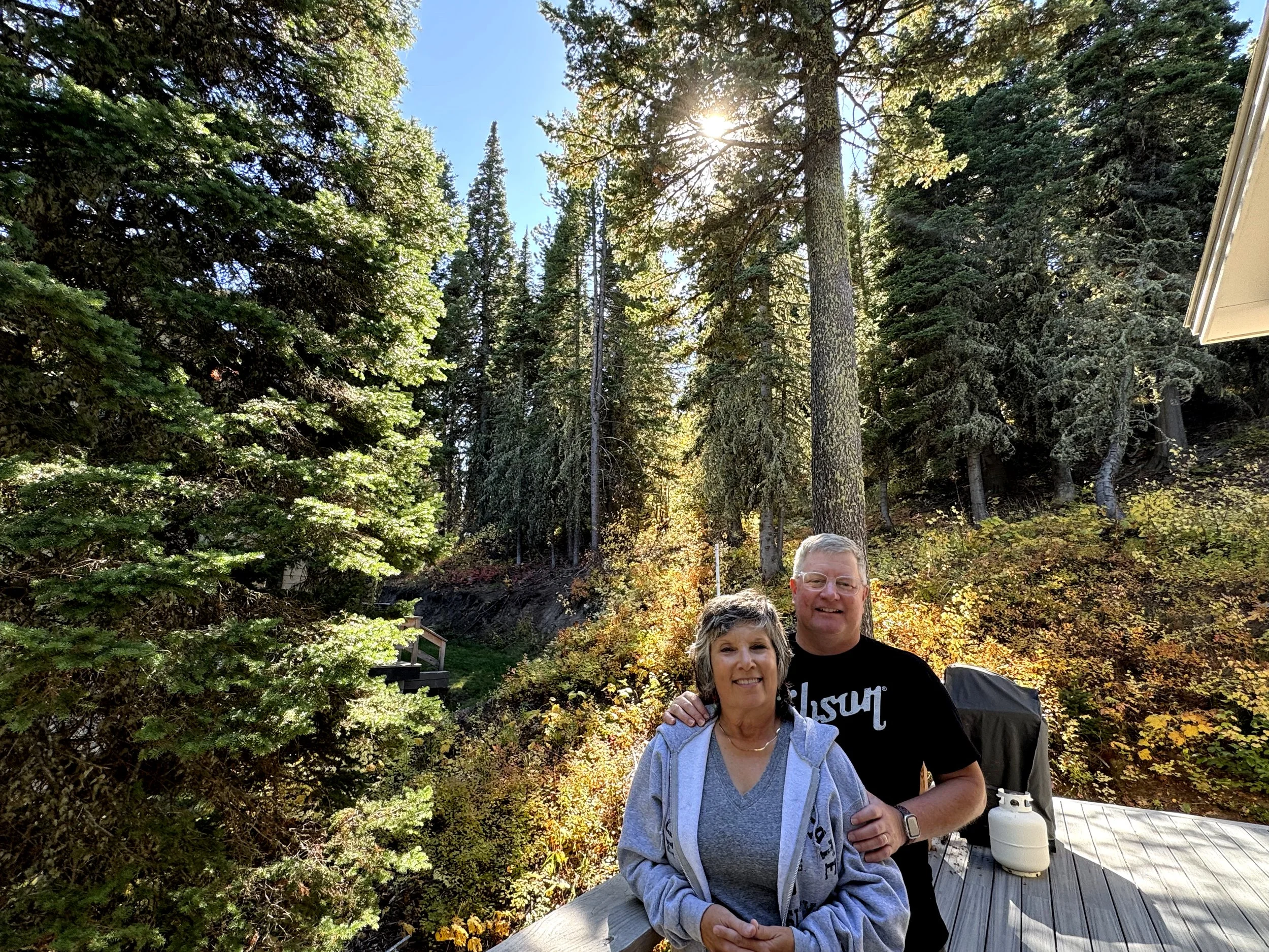 A smiling woman with short gray hair and a man with glasses and short gray hair standing together outdoors in a wooded area with tall trees and sunlight filtering through the branches.