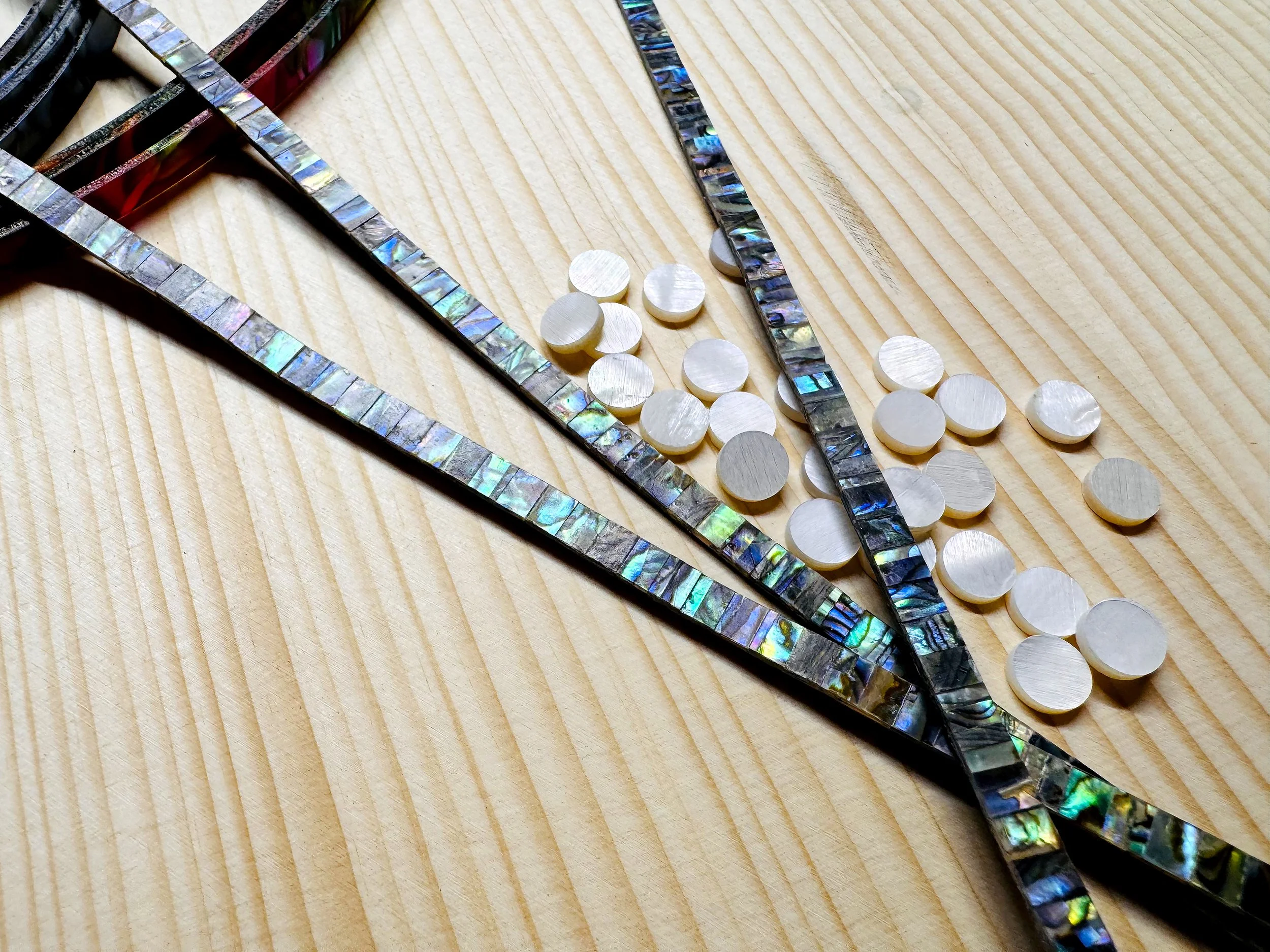 Close-up of abalone inlay or mosaic pieces, including iridescent strips and circular shell pieces, arranged on a wooden surface.