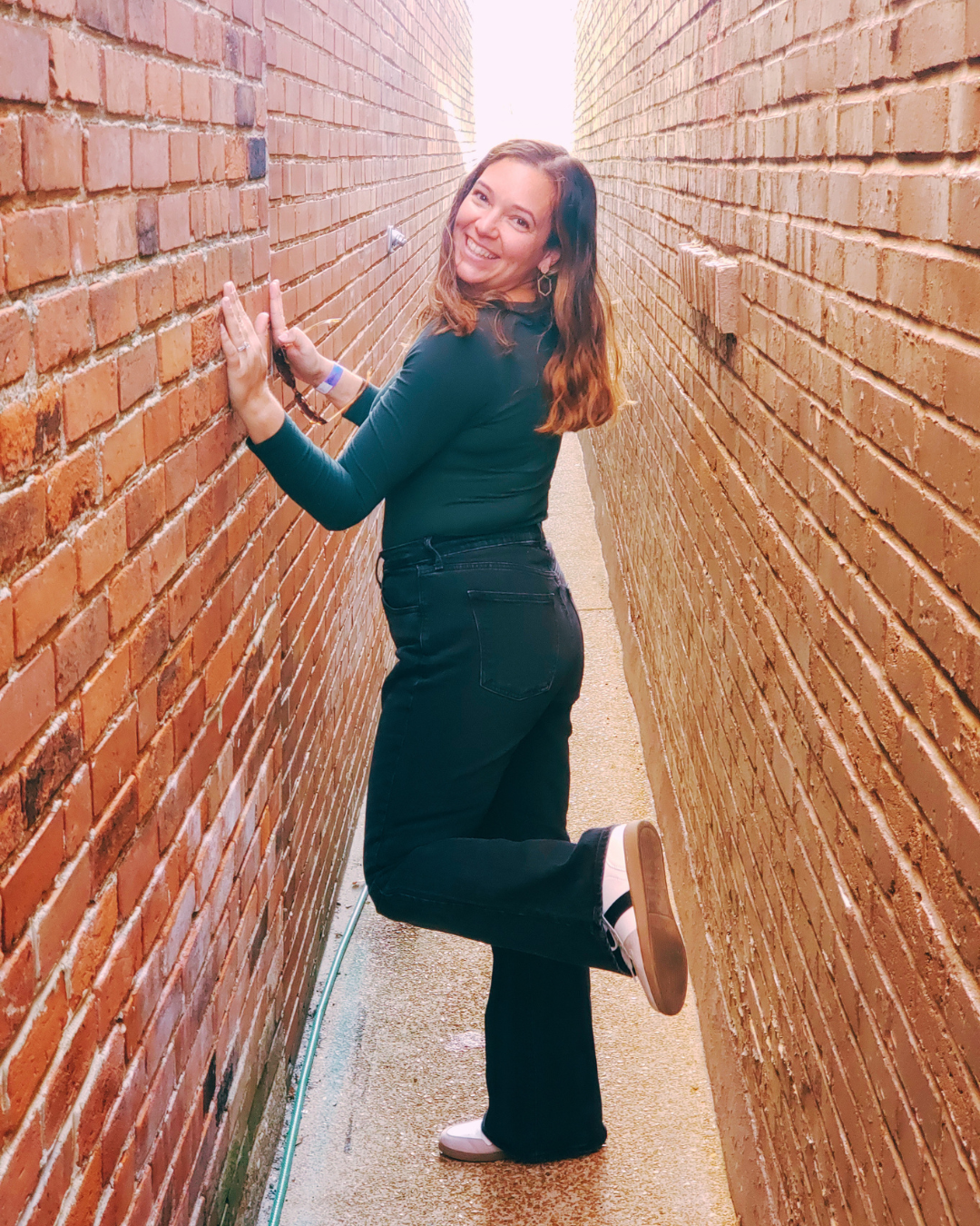 A young woman with long brown hair, wearing a blue long-sleeve shirt, black jeans, and sneakers, smiling and posing in a narrow alleyway between two brick walls.