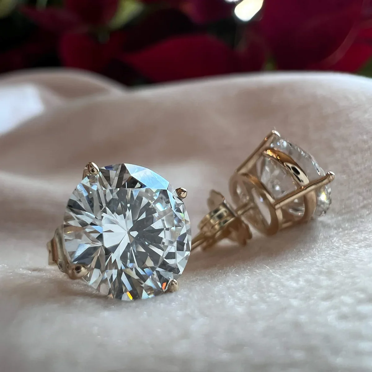 A pair of diamond stud earrings with large round-cut diamonds set in yellow gold, placed on a soft white fabric surface.