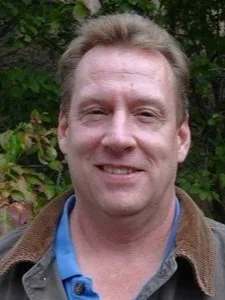 A man with short light brown hair smiling, wearing a brown jacket over a blue shirt, outdoors with green foliage in the background.