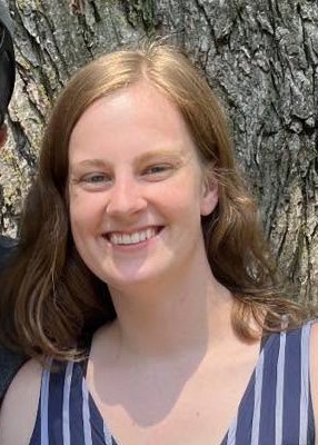 A young woman with light red hair and a big smile, wearing a sleeveless, striped blue and white top, standing outdoors in front of a large tree trunk.