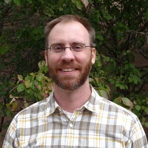 A man with glasses and a beard smiling outdoors with green foliage in the background.