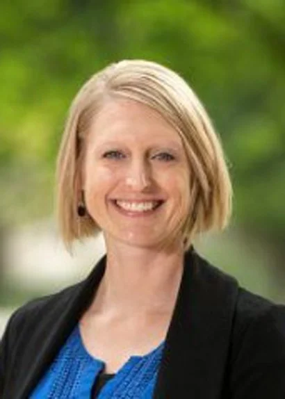 A woman with blonde, shoulder-length hair smiling outdoors with a blurred green background.
