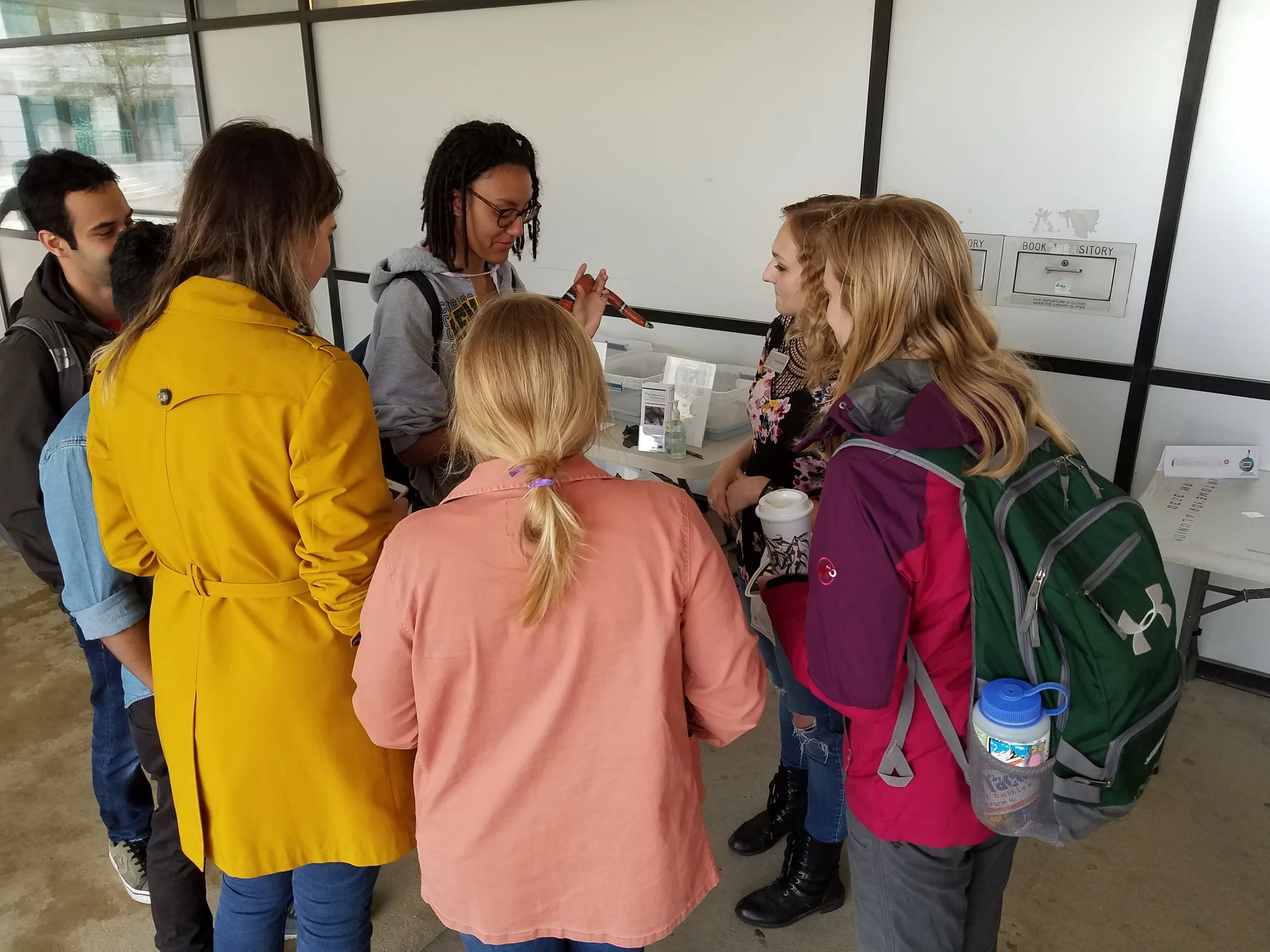Group of six people gathered around a table with various items, engaged in conversation inside a building with large windows.