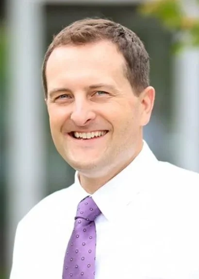 Portrait of a smiling man wearing a white shirt and purple tie, outdoors with blurred greenery in the background.