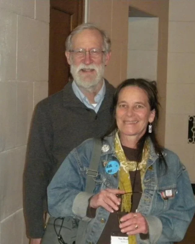 A smiling man with glasses and a beard standing behind a woman with dark hair, wearing a denim jacket and a yellow scarf, inside a building with brick and tile walls.