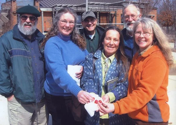 Six people standing outside, in front of a building, smiling, with some wearing glasses and jackets. Two women are holding a small red object together, and one woman is holding a small notebook or planner.