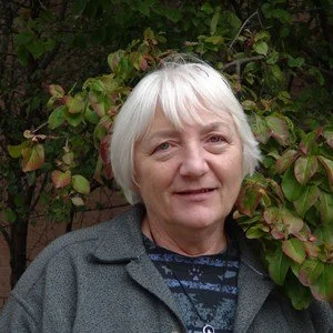 An elderly woman with short white hair, wearing a gray coat, standing outdoors in front of green foliage.