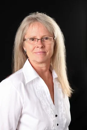 A woman with long gray hair, glasses, and a white button-up shirt, standing against a black background.