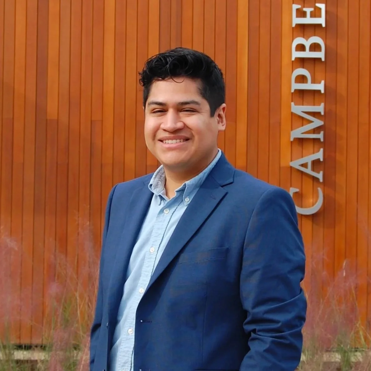 A man with dark hair wearing a blue blazer and light blue button-up shirt standing outdoors in front of a wooden wall with the word 'CAMPBELL' on it.