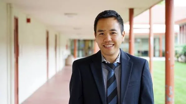 A smiling man in a dark suit and tie standing outdoors on a walkway with a building and green grass in the background.