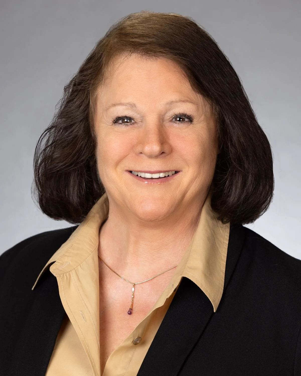 A professional headshot of a middle-aged woman with shoulder-length dark brown hair, smiling, wearing a beige collared shirt under a black blazer, and a delicate necklace, against a plain gray background.