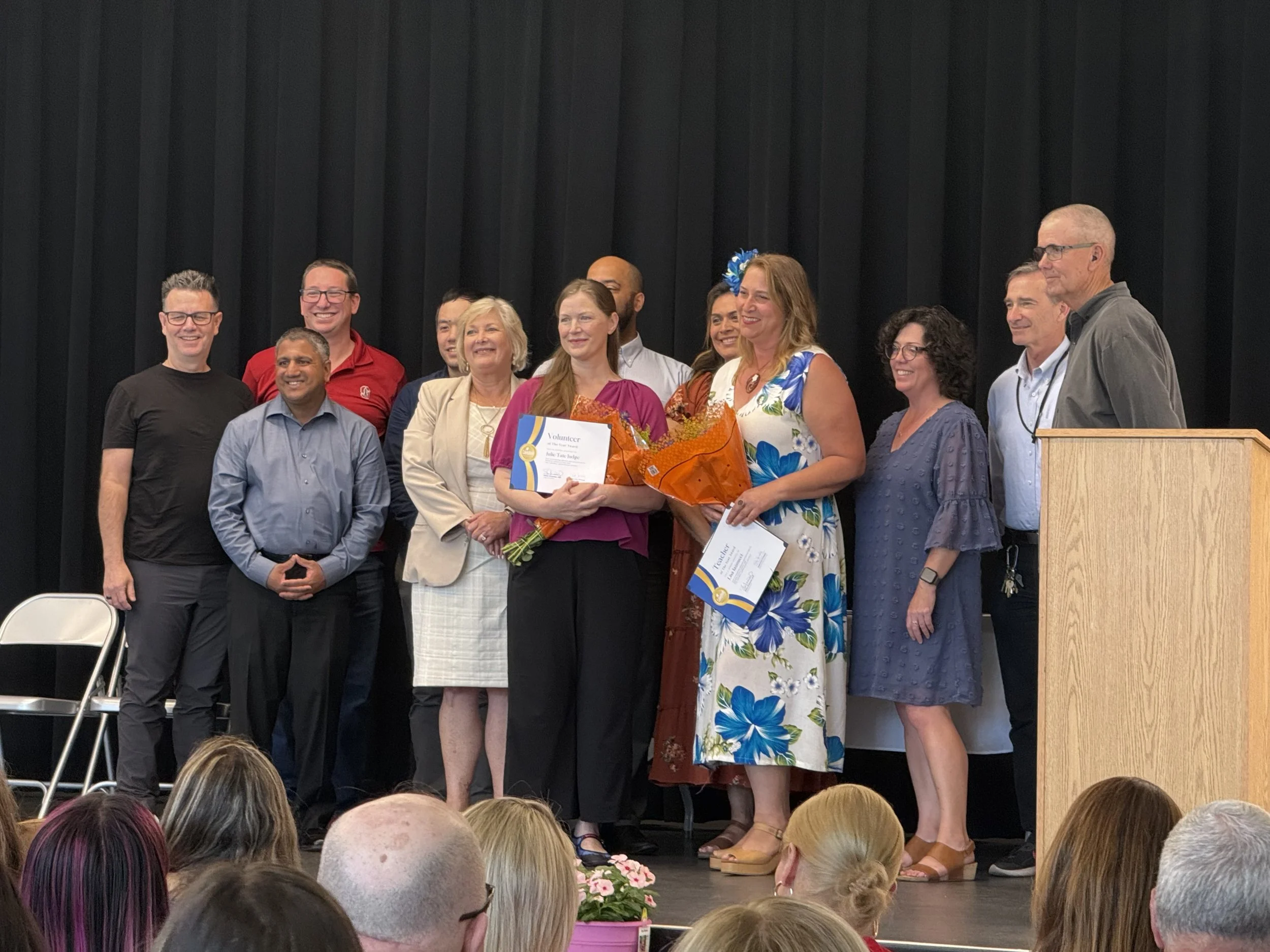 Group of diverse people on stage at an award ceremony, with one woman holding a bouquet of flowers and a certificate. Audience members are visible in the foreground.