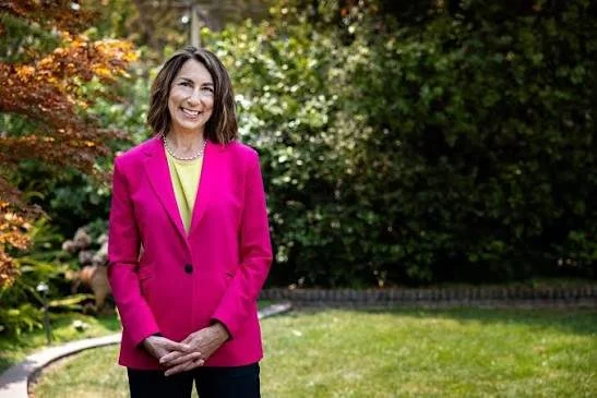 Woman with shoulder-length brown hair wearing a bright pink blazer and a yellow top, standing outdoors in a garden with green trees and plants.