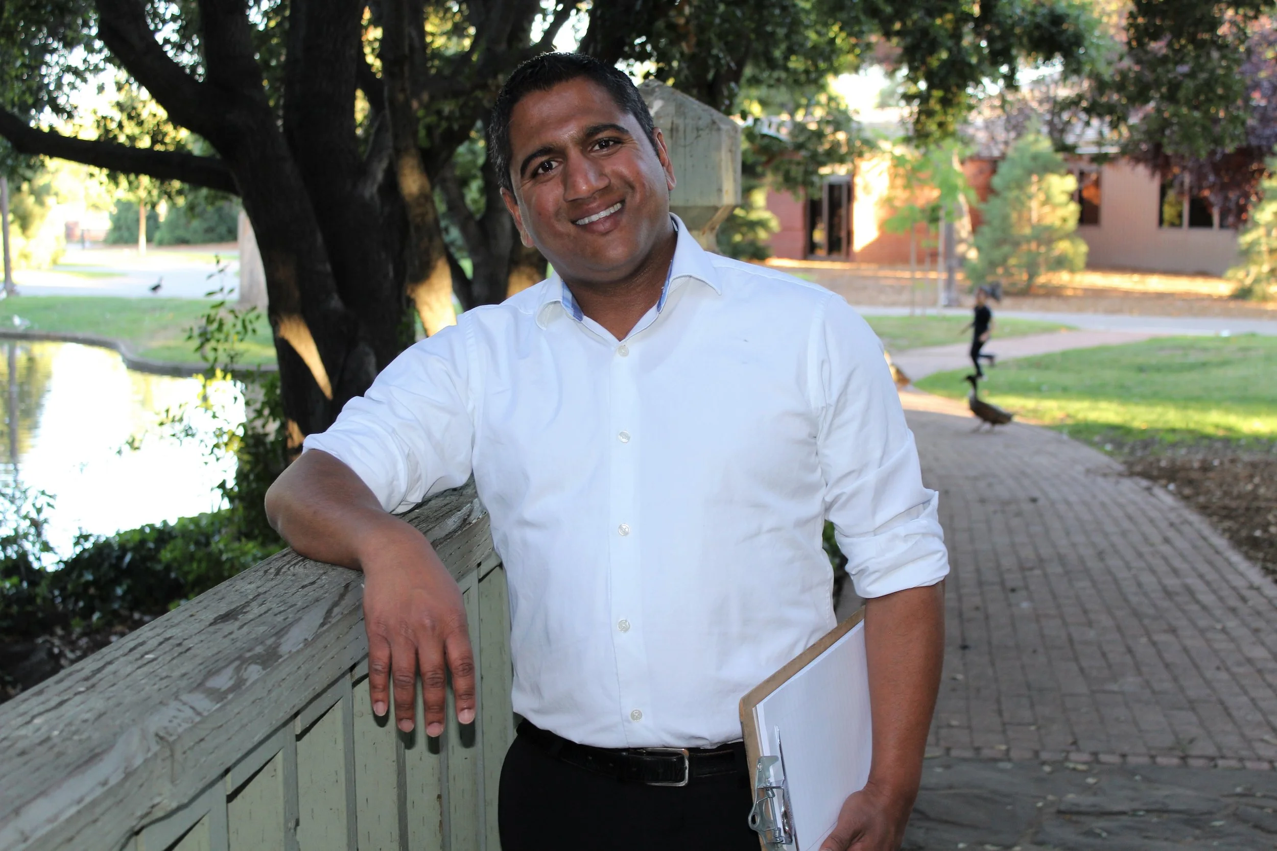A man with dark hair and a white dress shirt smiling outdoors, leaning on a wooden railing near a pond, with trees and a brick building in the background in the late afternoon.