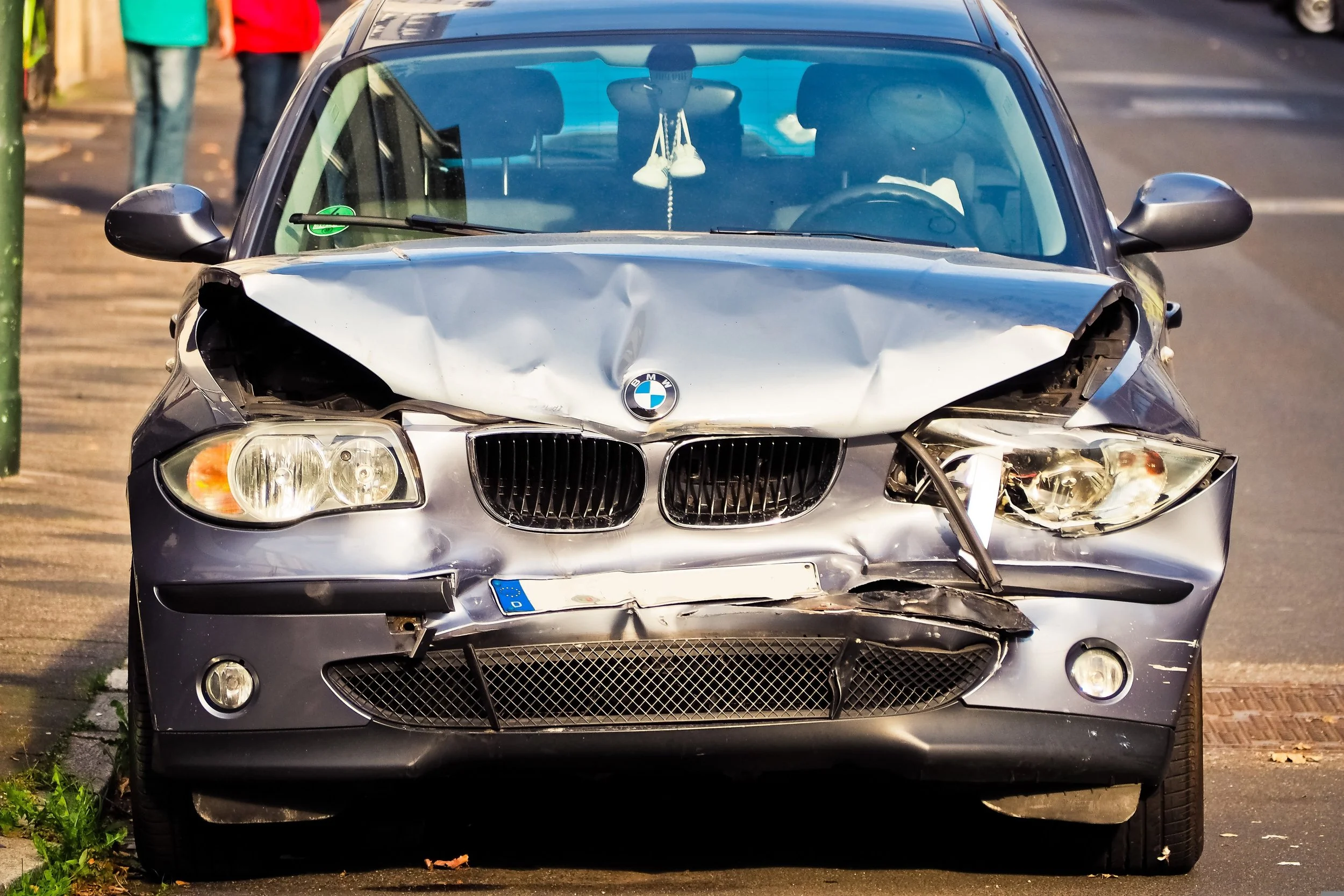Front view of a silver BMW car with front-end damage, parked on the street. The hood is crumpled, and the front bumper is cracked. The right headlight is damaged, and the left headlight appears intact. Inside the car, a pair of white shoes hang from the rearview mirror.