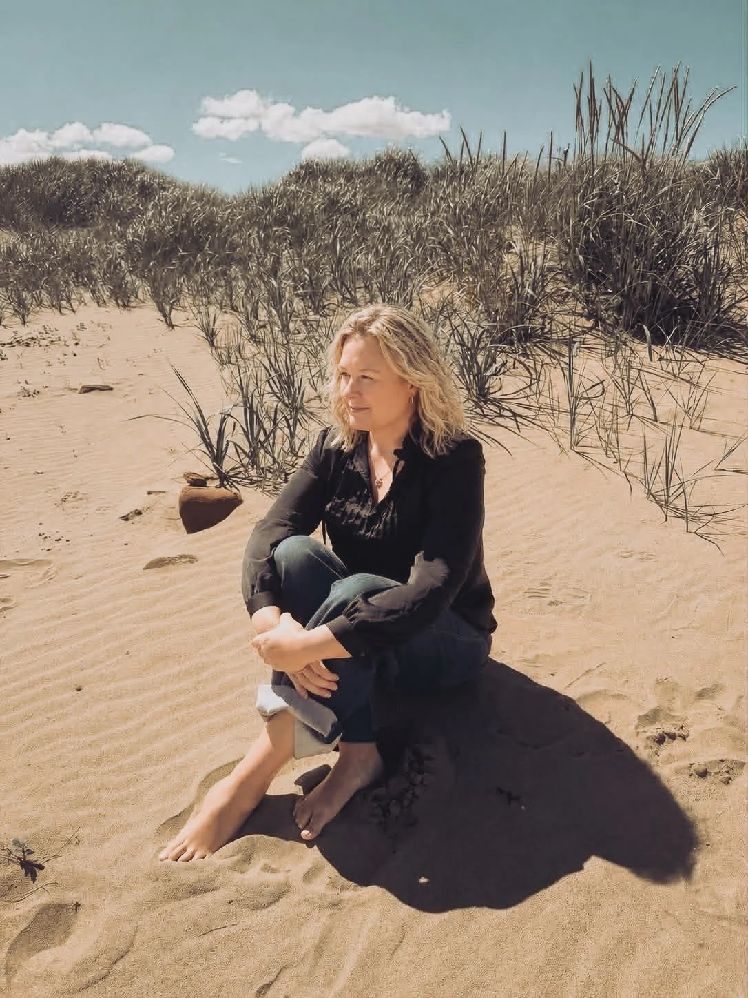 A woman with blonde hair sitting barefoot on sand with her arms wrapped around her knees in a beach setting, with sand dunes and tall grass behind her and a partly cloudy sky above.