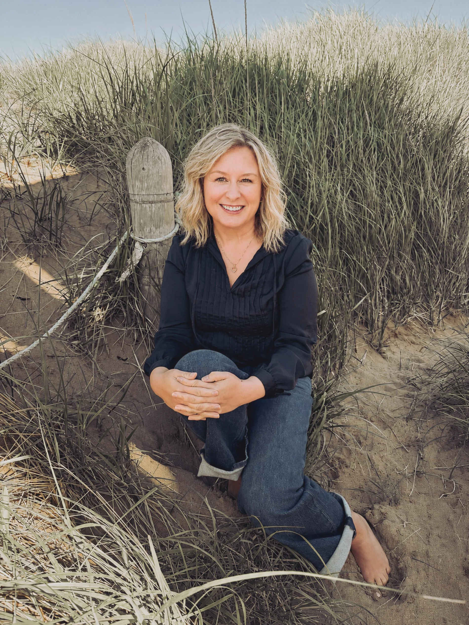 A woman with blonde curly hair, wearing a black shirt and blue jeans, sits on sandy ground in front of dune grass and wooden posts at beach, smiling at camera.
