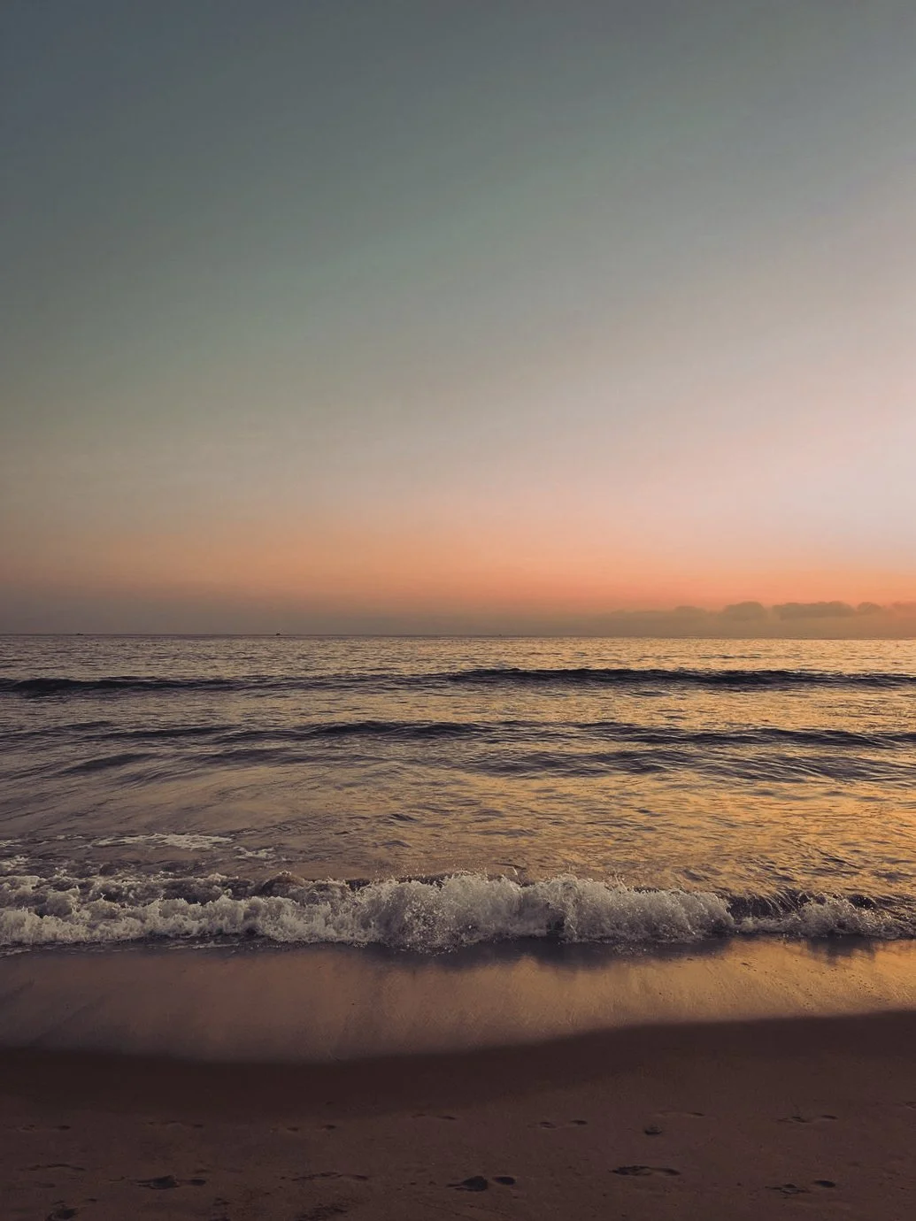 Sunset over the ocean with gentle waves lapping the sandy beach.