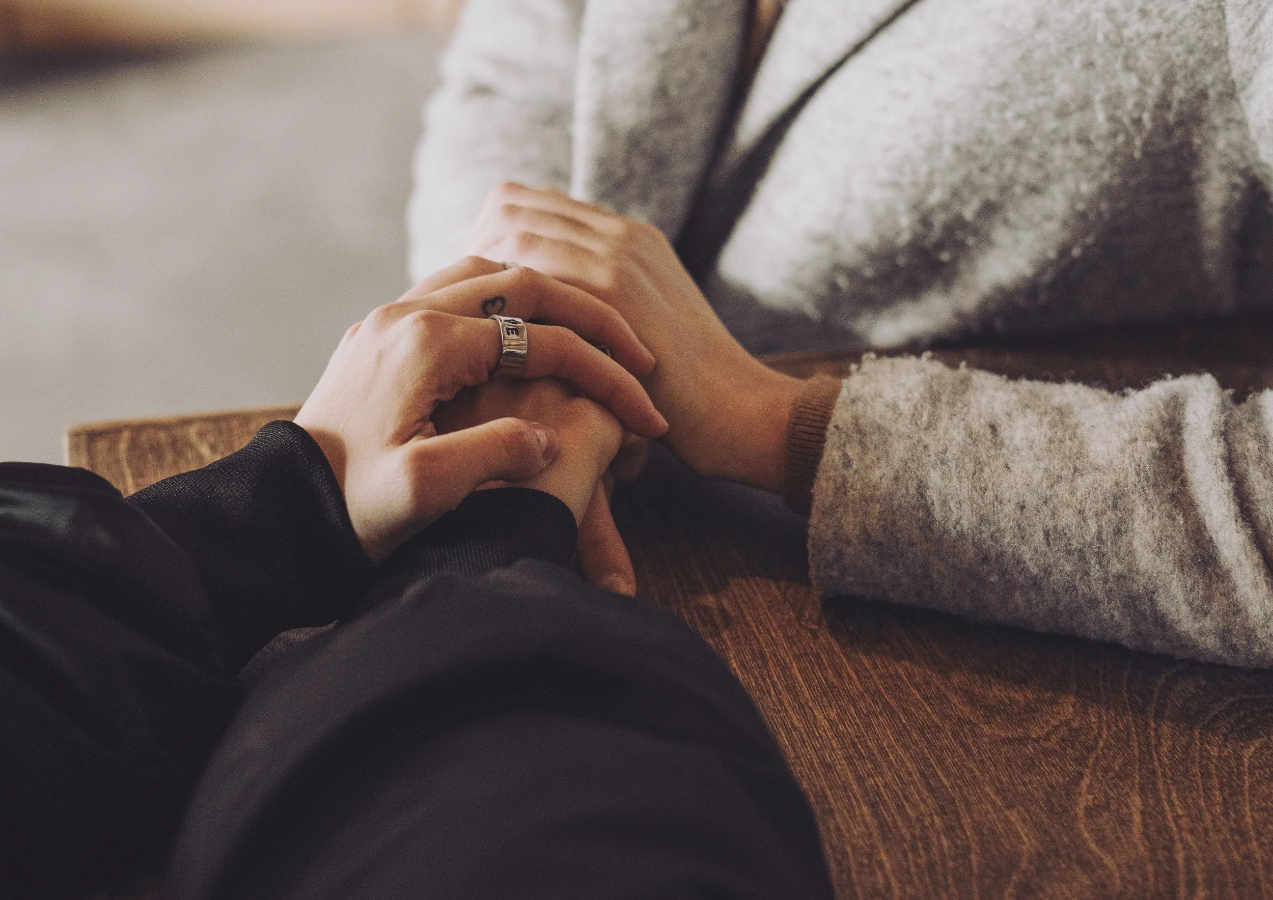 Close-up of two people holding hands on a wooden table, with one person wearing a ring with the word 'LOVE' and a small heart tattoo visible.